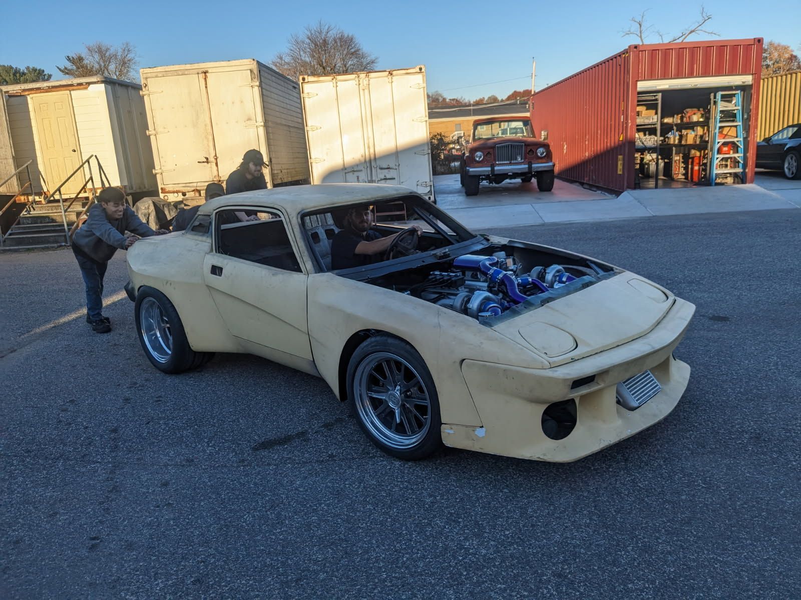 Tan custom car with open hood being pushed by a person, others nearby.