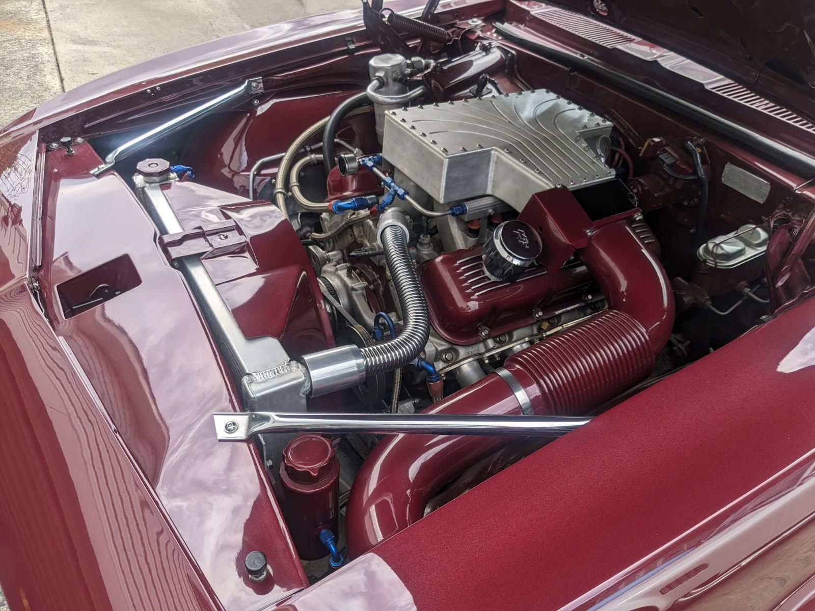 Maroon car engine bay, custom components, polished metal, tubular intake, and bright lighting.