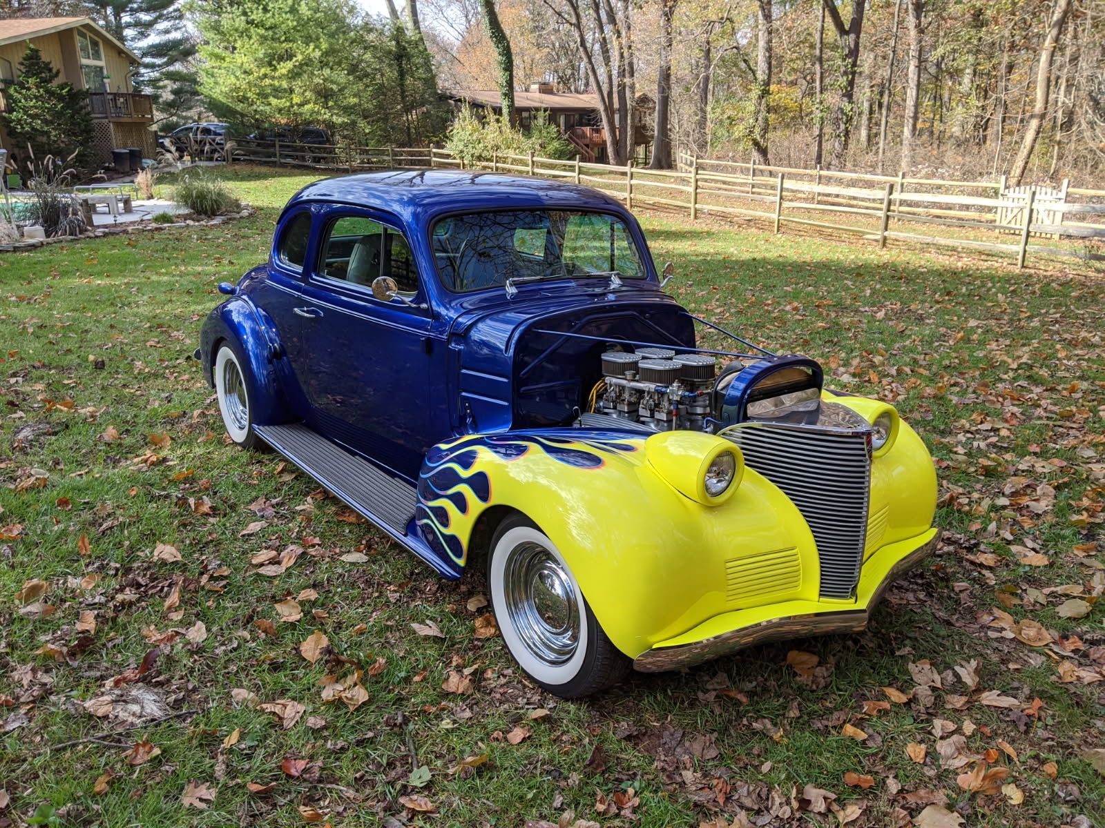 Blue and yellow hot rod car with flames in a yard with fall leaves.