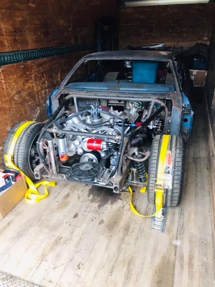 Blue car chassis with engine and roll cage in a truck, secured with yellow straps.