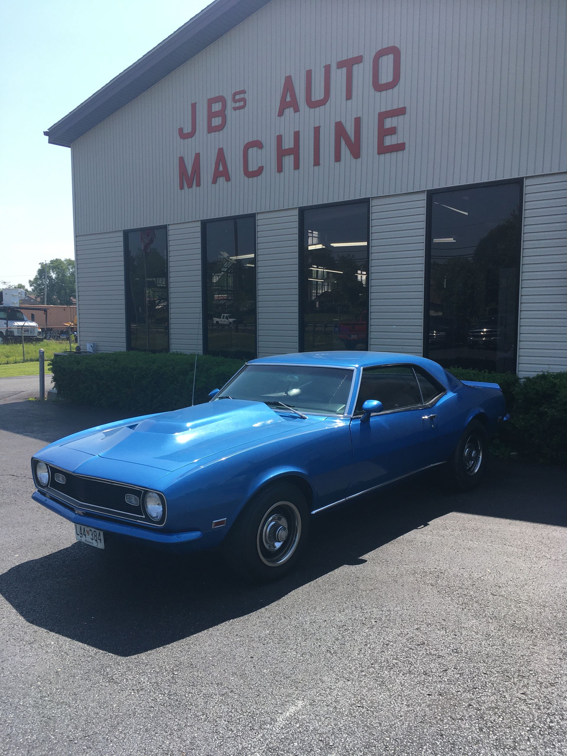 Blue classic car parked in front of JB's Auto Machine. Building is white with red letters.