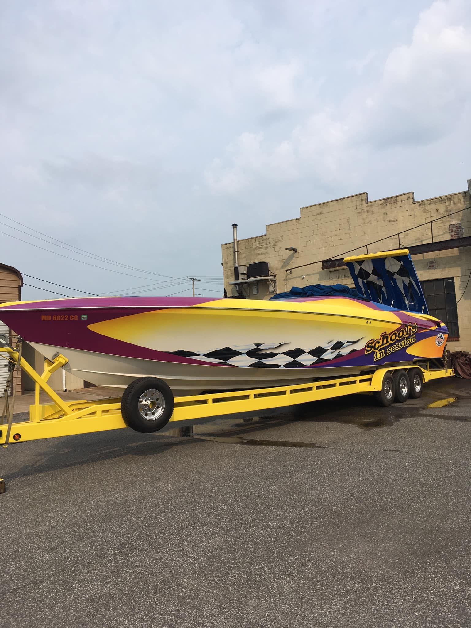 Speed boat on a yellow trailer, multi-colored design with purple and yellow accents parked outside a building.