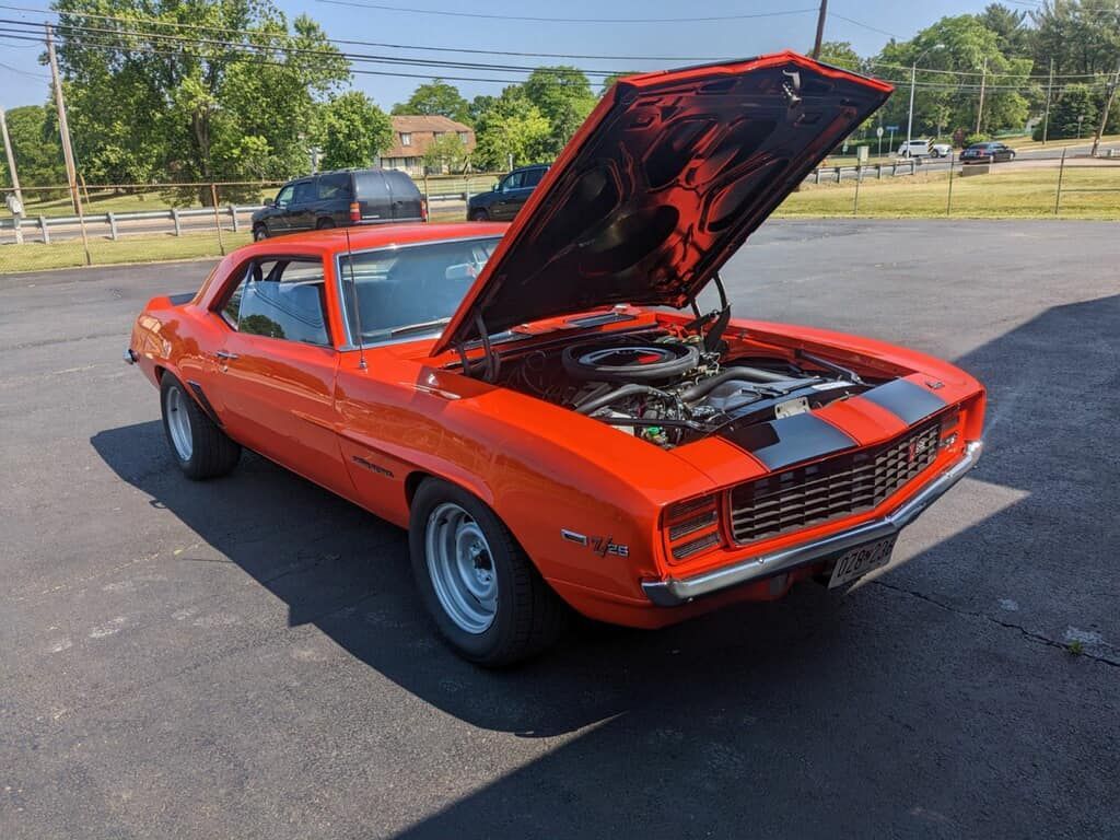 Orange classic muscle car with black stripes, hood open, on asphalt in front of a building.