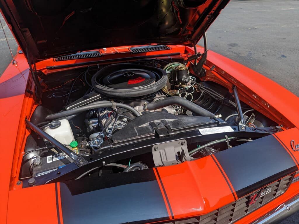 Bright orange classic Camaro with black racing stripes, hood open showing engine.