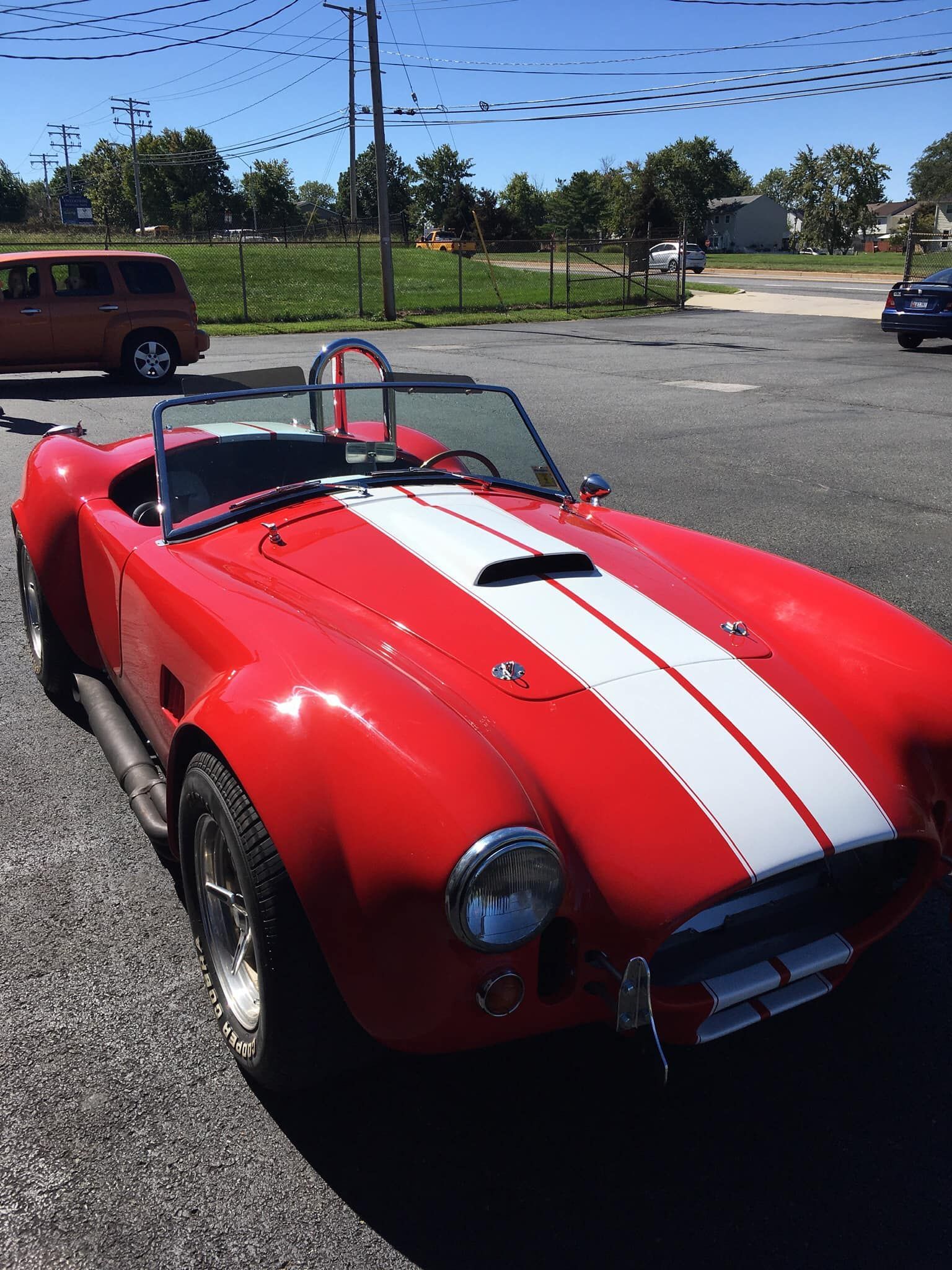 Red Shelby Cobra sports car with white racing stripes, parked outdoors.