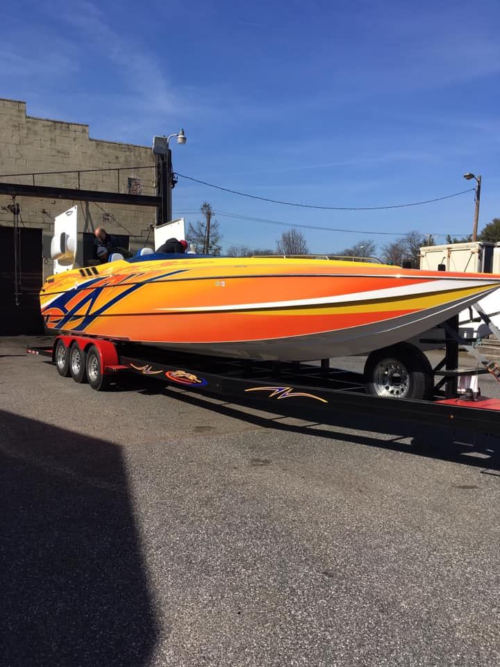 Orange and yellow speed boat on a trailer, parked on pavement under a blue sky.
