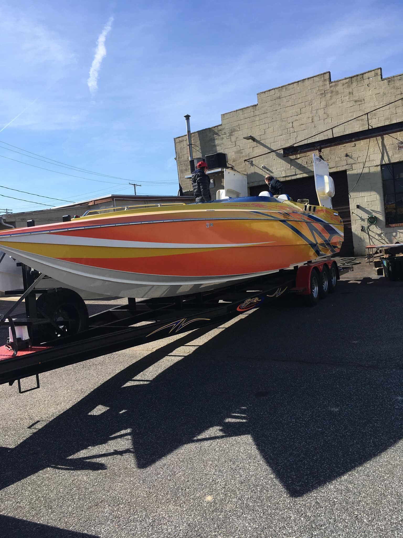 Orange and yellow speedboat on a trailer, parked outside a brick building on a sunny day.