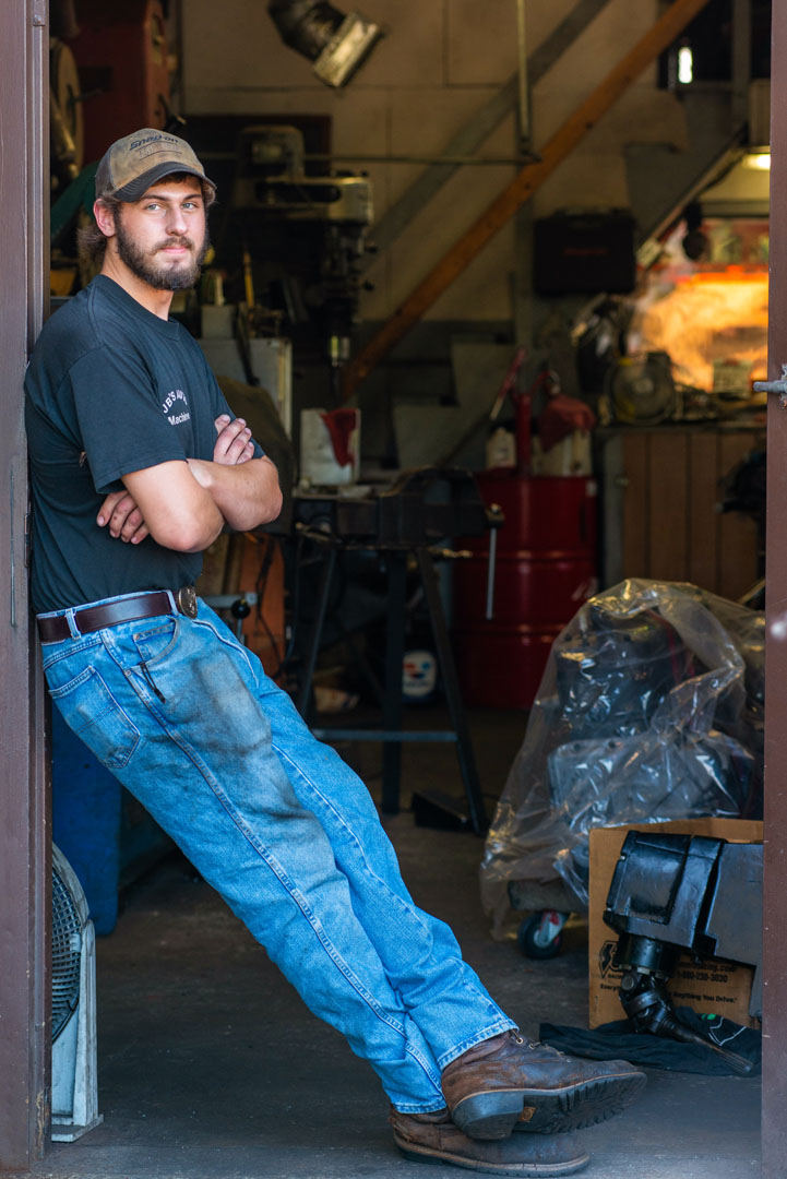 Man in work clothes leans against a doorway in a workshop, arms crossed, wearing a cap.