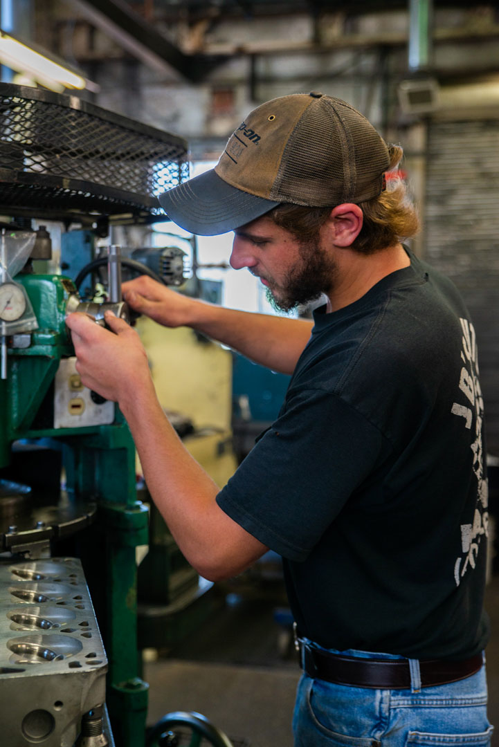 Mechanic in cap adjusting a machine in a workshop.  He is wearing jeans and a black shirt.
