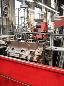 Engine block being worked on in a machine shop. A person in a red shirt stands near the machinery.