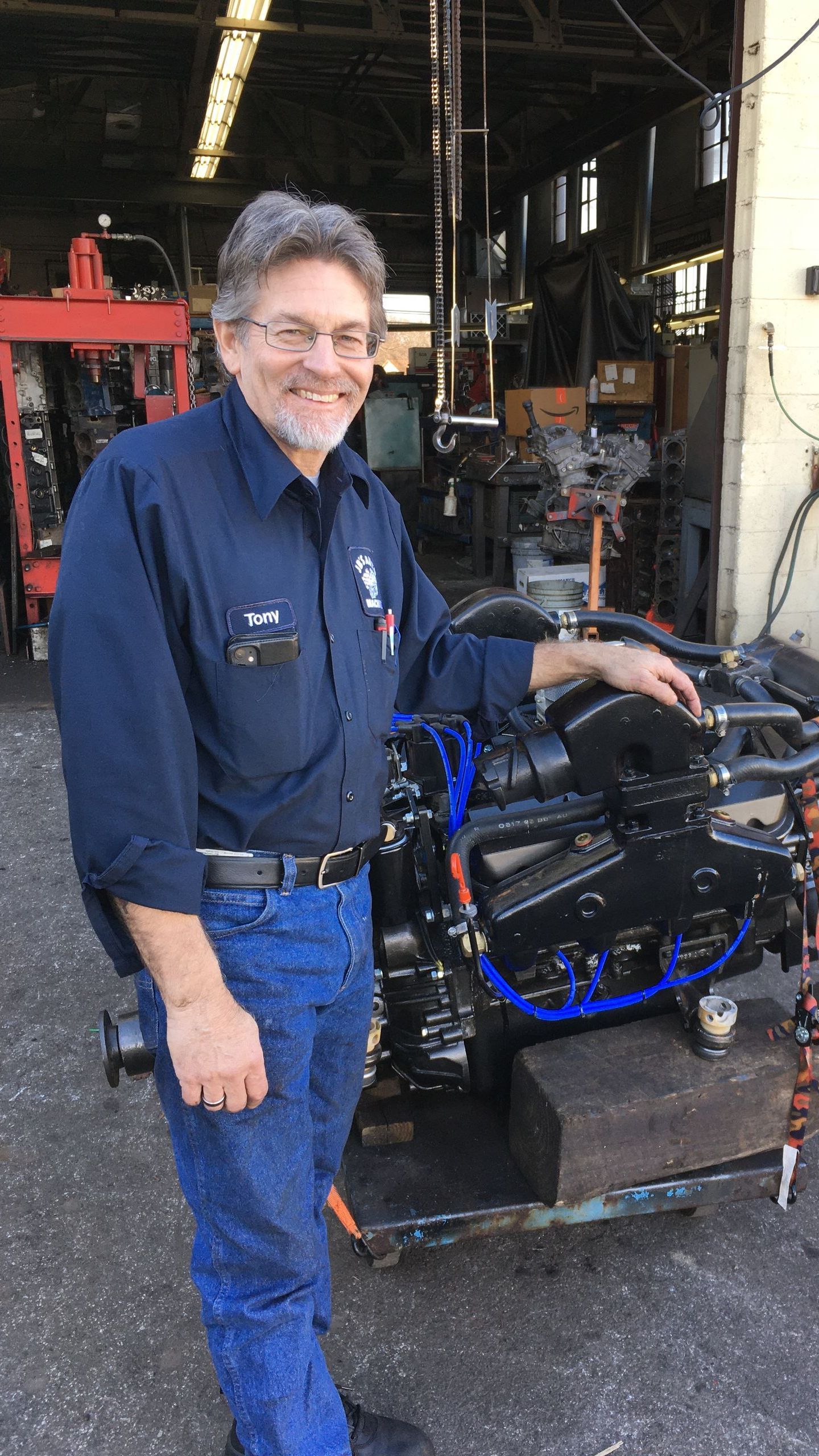 Man in blue work shirt smiles beside a black engine, likely in a workshop.