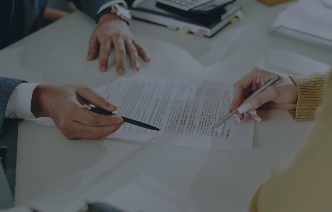 Hands pointing at and signing a document on a white table. A calculator and notepad are in the background.
