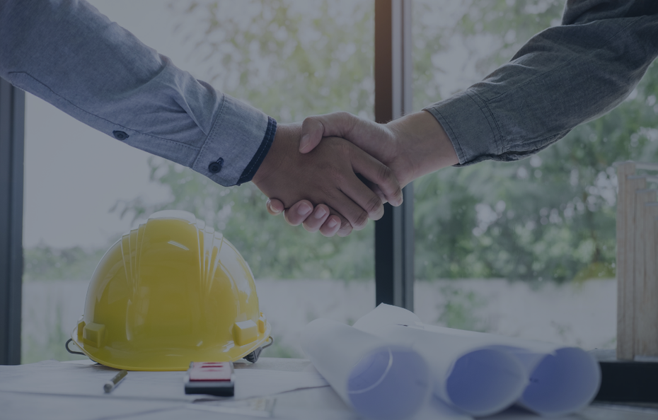 Two people shaking hands over a table with blueprints, a yellow hard hat, and a window in the background.