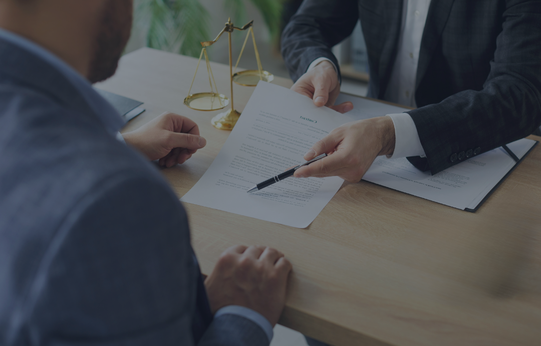 Person in suit pointing at document, sitting across from another person at a desk. Legal scales are in the background.