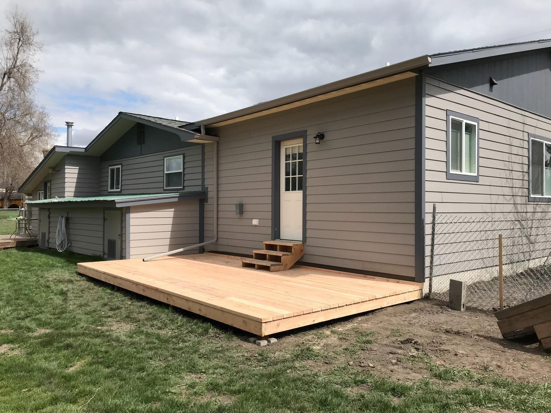 Wooden deck extending from a gray house with a door and steps. Green grass and blue sky are visible.