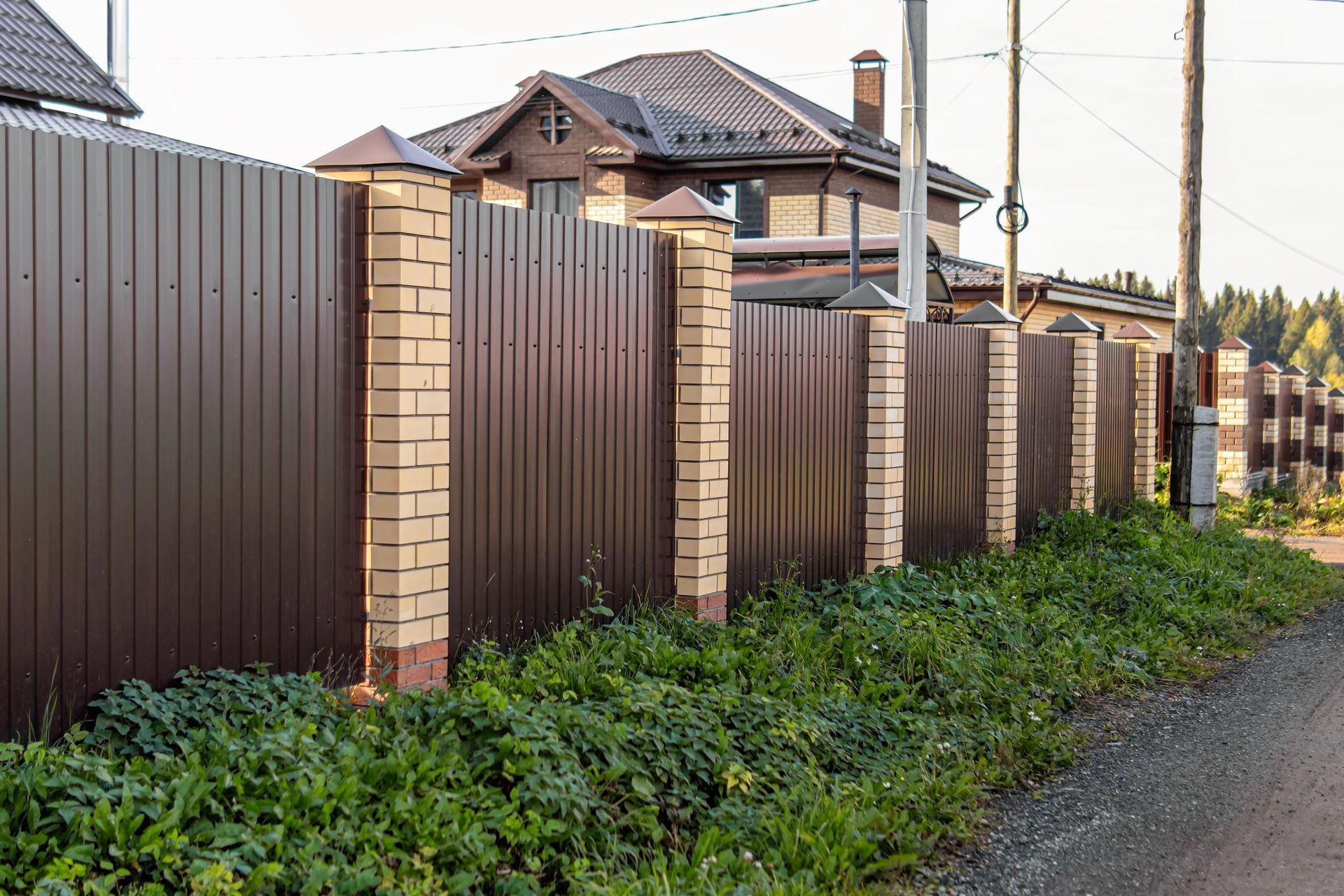 Brown metal fence with brick pillars bordering a house and road.
