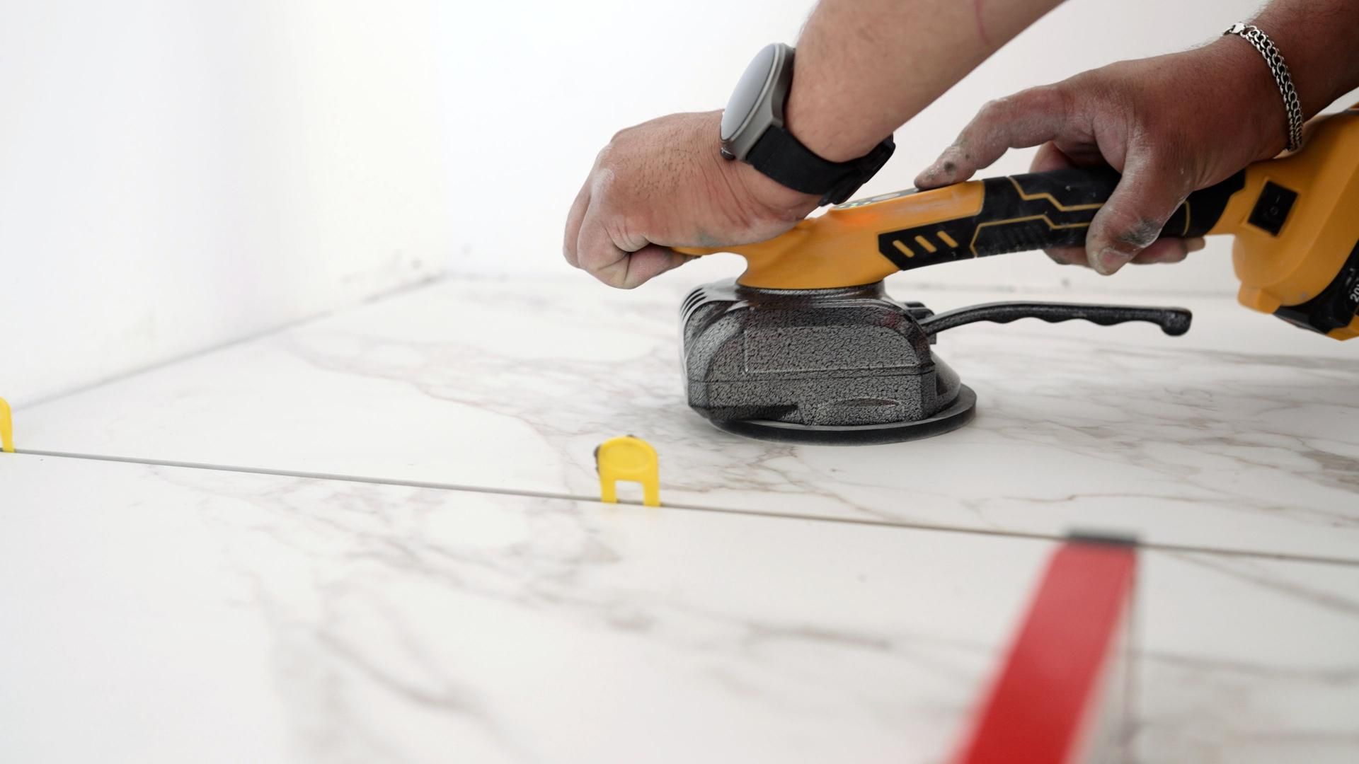 A person uses a yellow tile-leveling tool on a marble tile floor, with tile spacers visible.