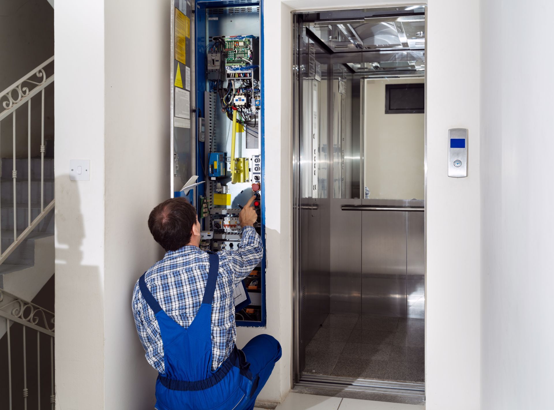 Technician in blue overalls repairing elevator controls with the doors open