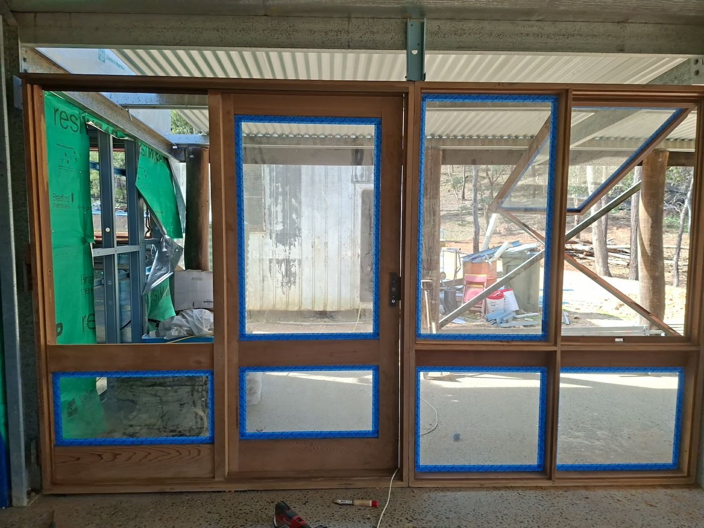 Wooden-framed windows with blue-taped panes inside a construction site overlook machinery and debris outside - Matty Stewart Painting & Decorating in Mudgee, NSW