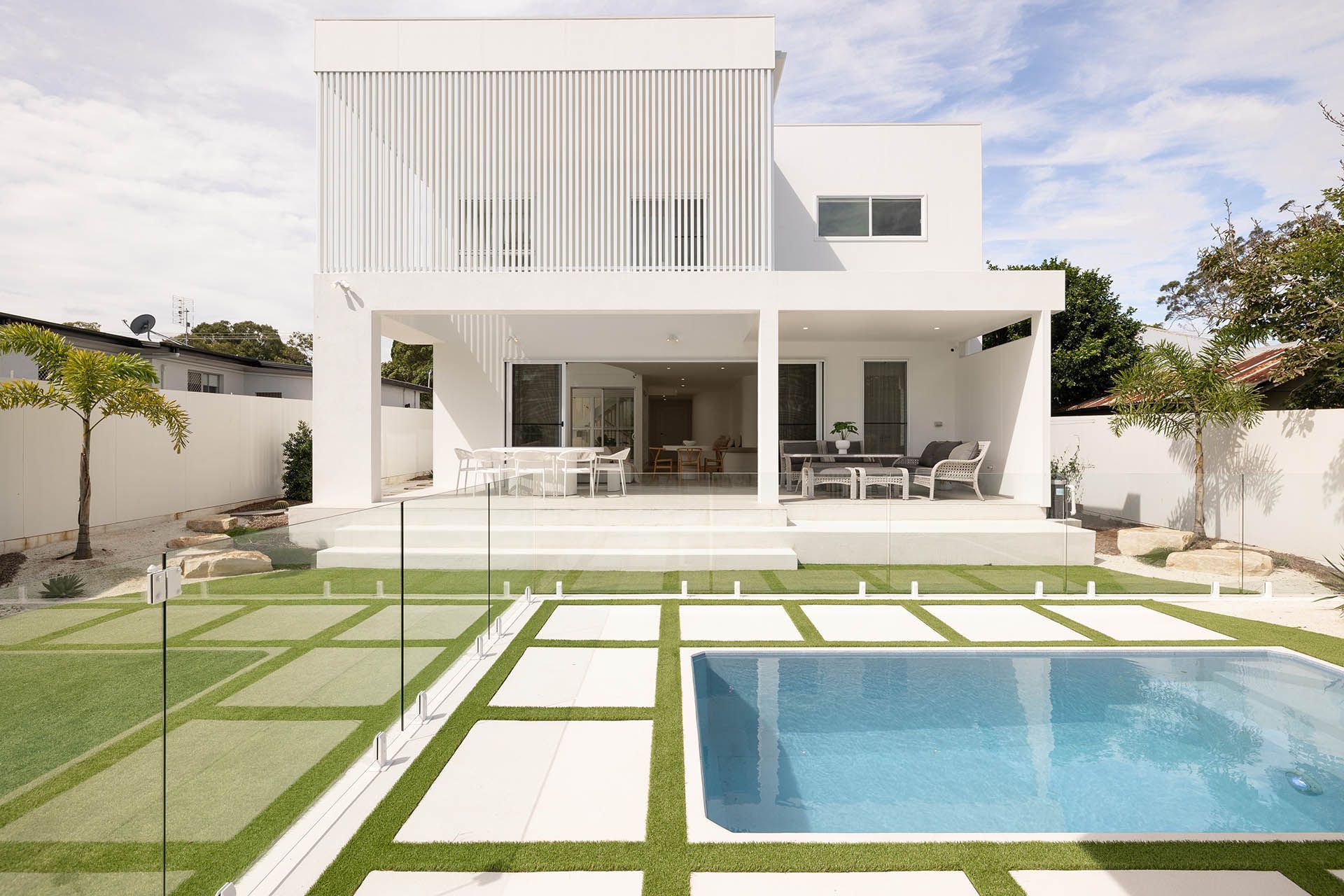 Modern white house with patio and lawn, viewed from a backyard pool and stone walkway