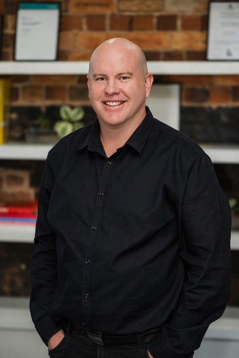 Smiling man in a black shirt standing indoors against a brick wall