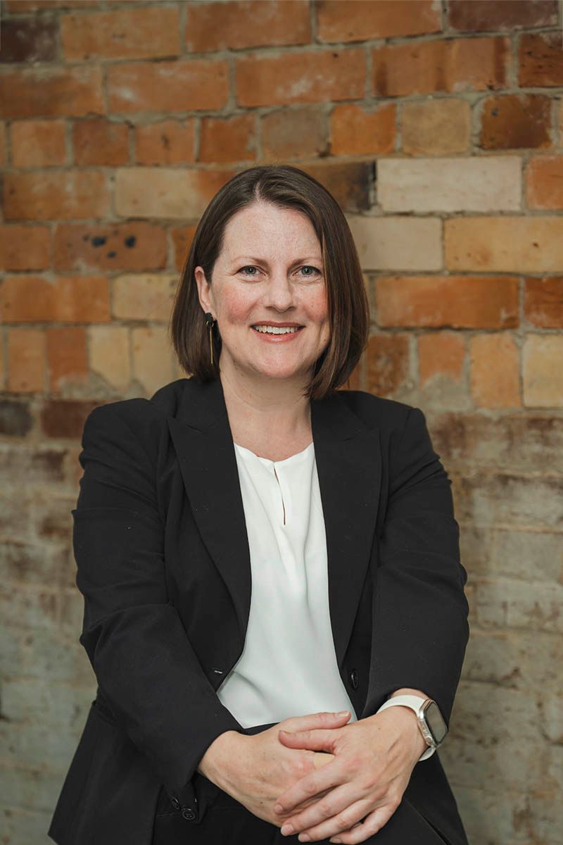 Professional headshot of a person in a black blazer and white blouse against a dark wood wall.