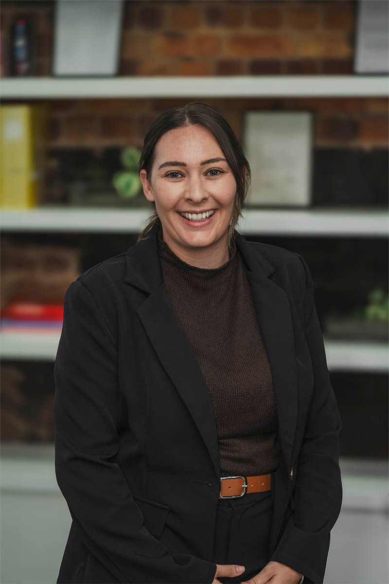 Smiling woman in a black blazer standing indoors before blurred shelves and books