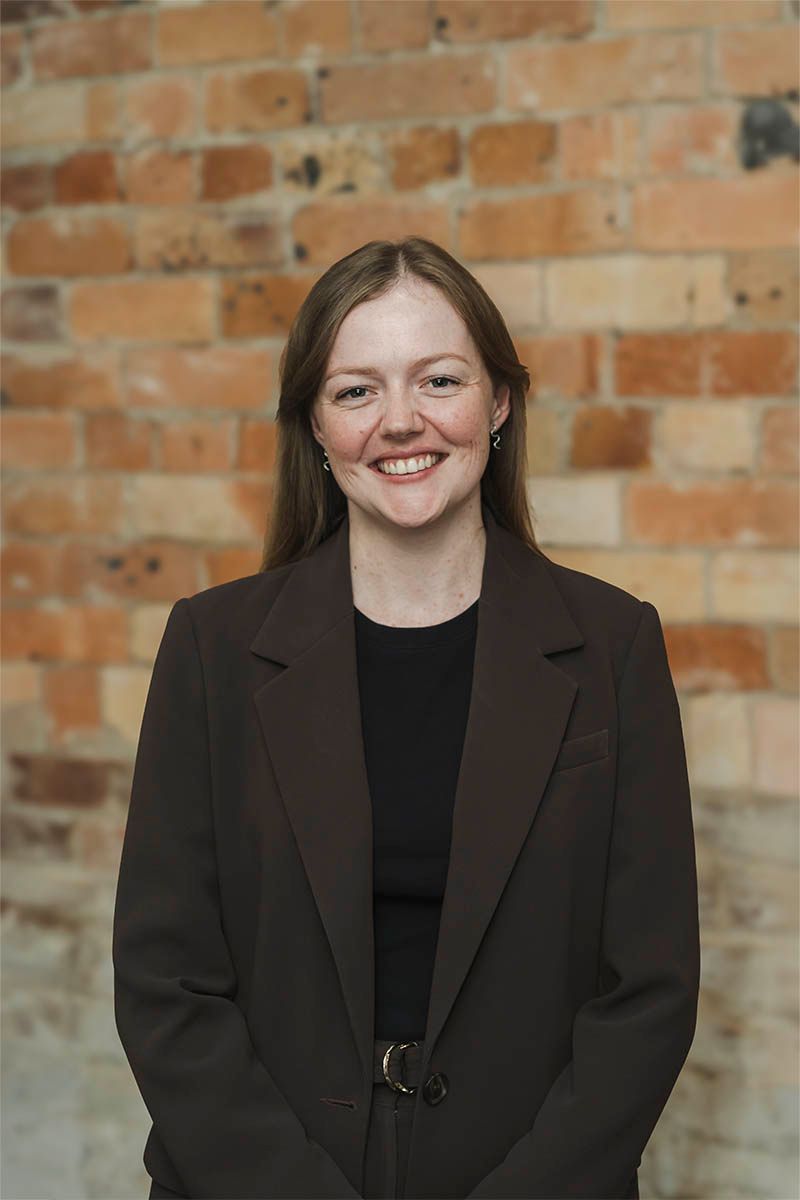 Woman in a dark blazer standing and smiling in front of a brick wall.