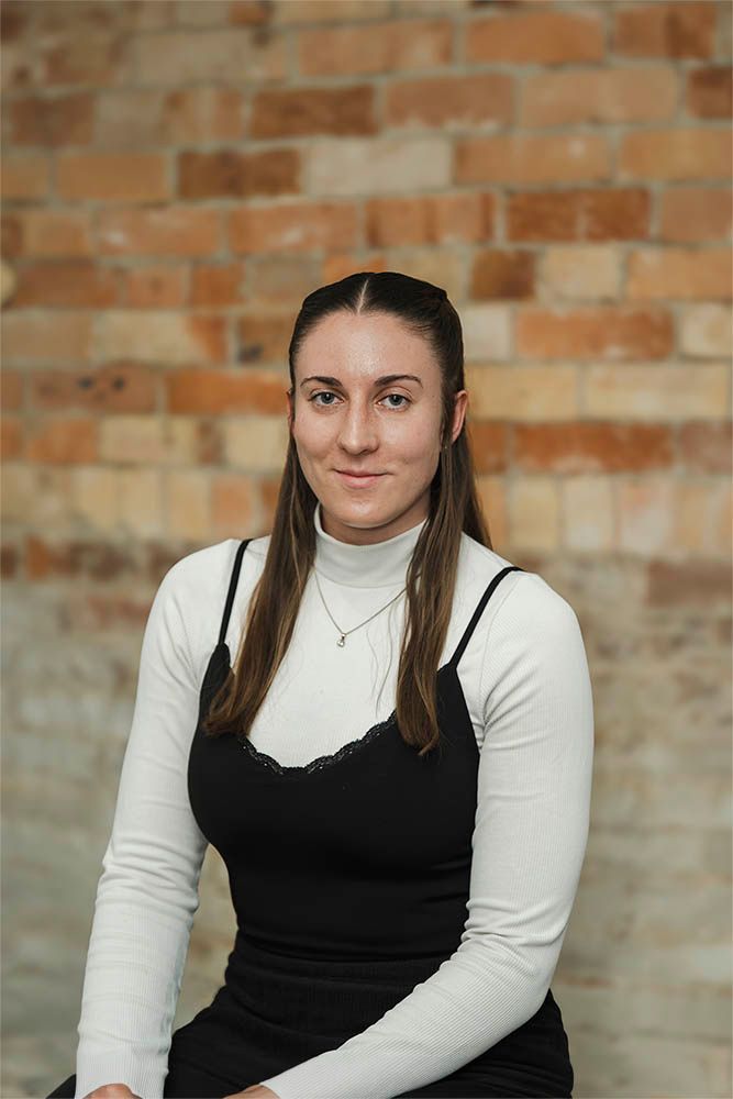 Seated woman in a white turtleneck and black dress against a brick wall, looking at the camera.