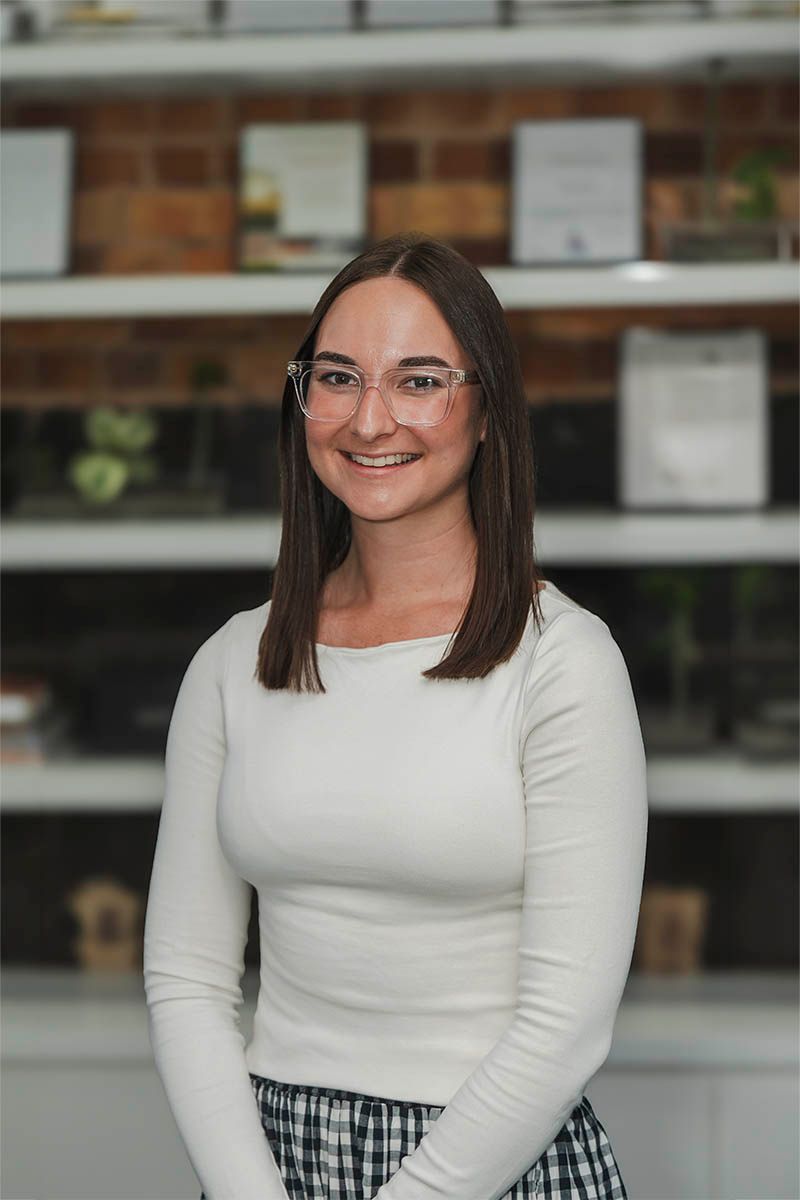 Smiling person in glasses and white top standing indoors in front of shelves