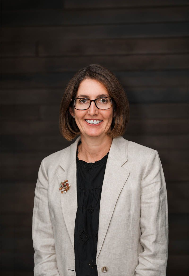 Smiling person in glasses and a light gray blazer against a dark wood backdrop