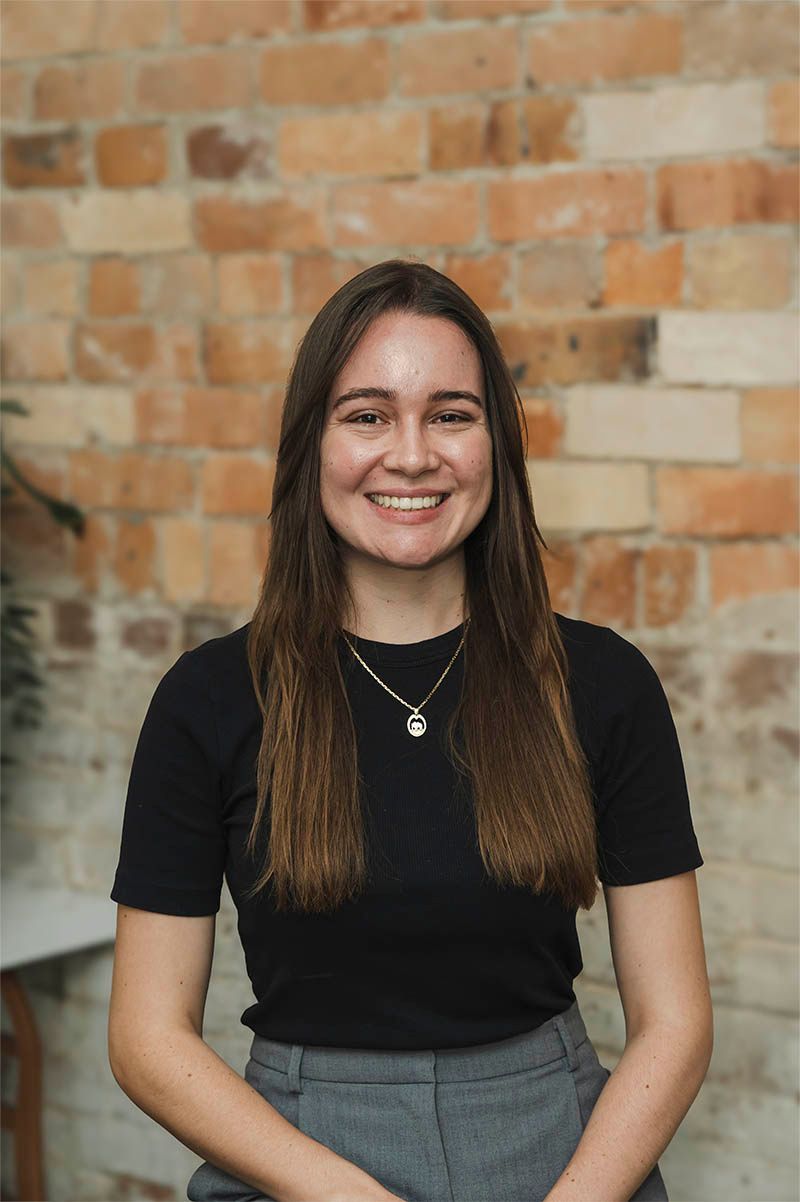 Smiling person in a black top and gray skirt seated against a brick wall.