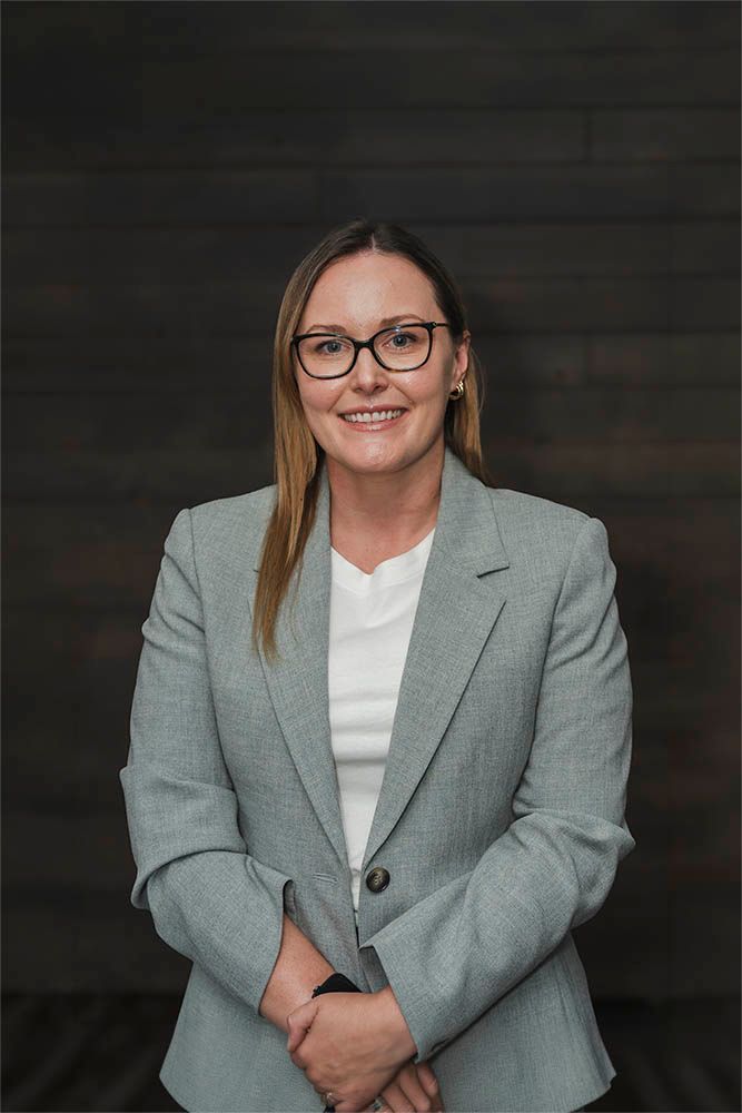 Smiling woman in a gray blazer and glasses standing against a dark wood wall.