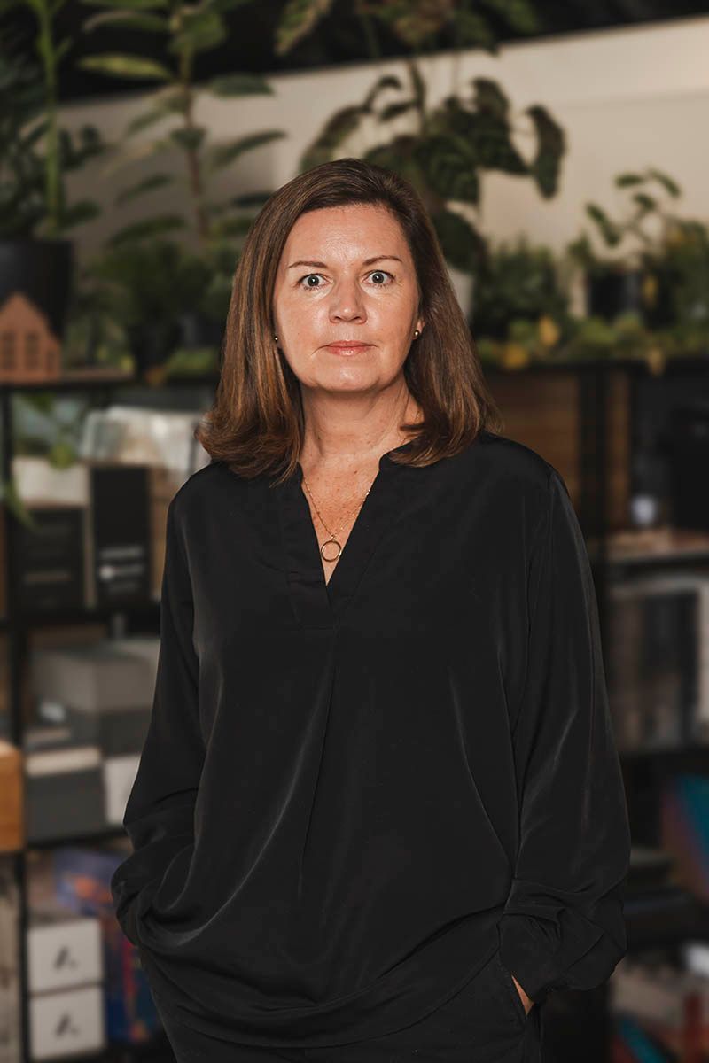Woman in a black blouse standing indoors with a blurred plant-filled background