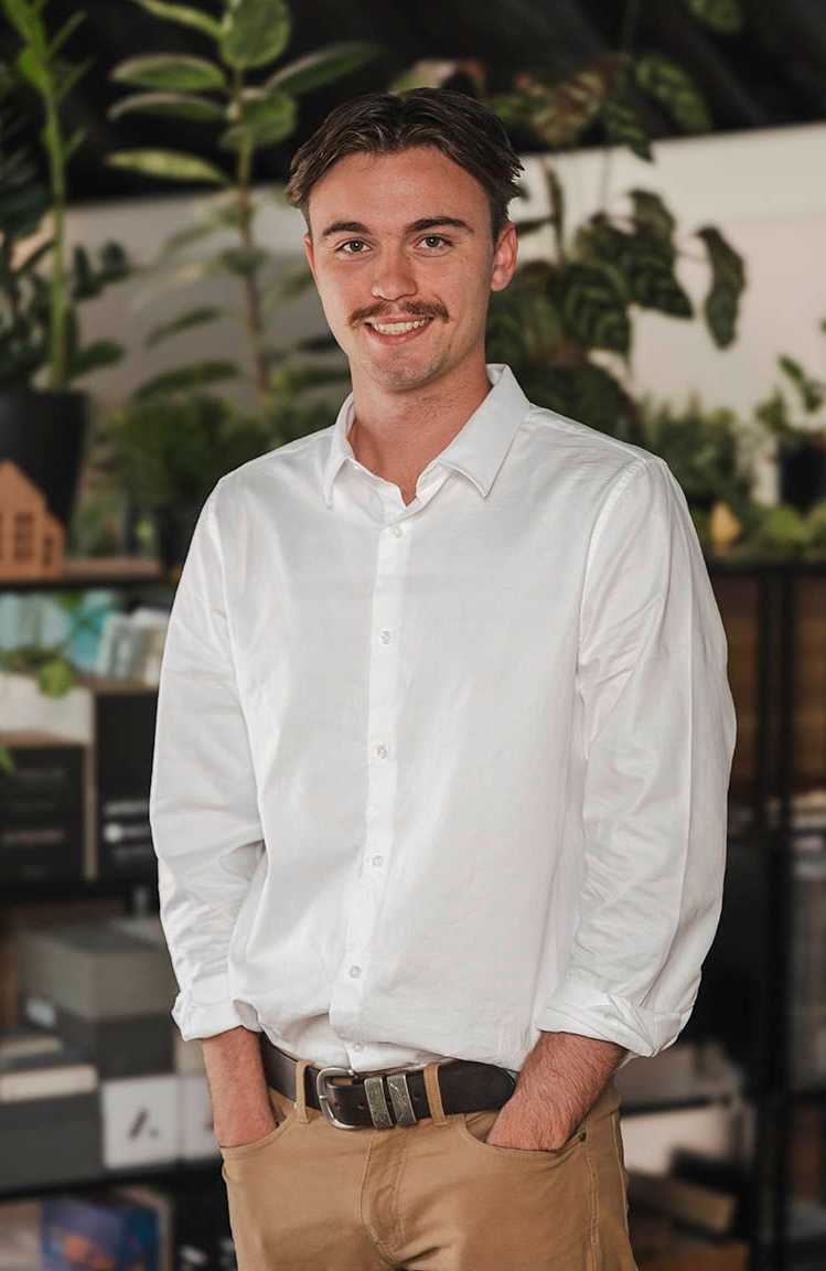 Man standing indoors in a white shirt and tan pants, smiling in front of shelves and plants.