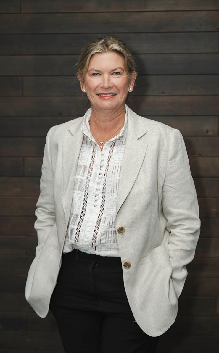 Woman standing against a dark wood wall, wearing a light blazer and white blouse, smiling confidently.
