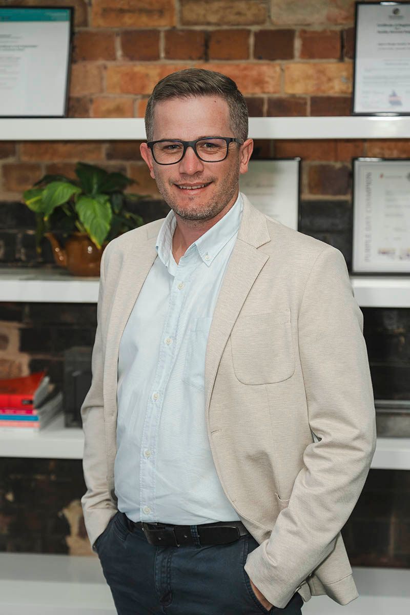 Man standing in a light blazer and glasses in front of shelves and a brick wall