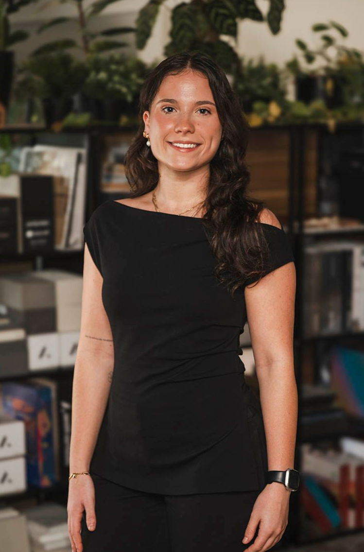 Smiling woman in a black dress standing indoors with shelves and plants in the background