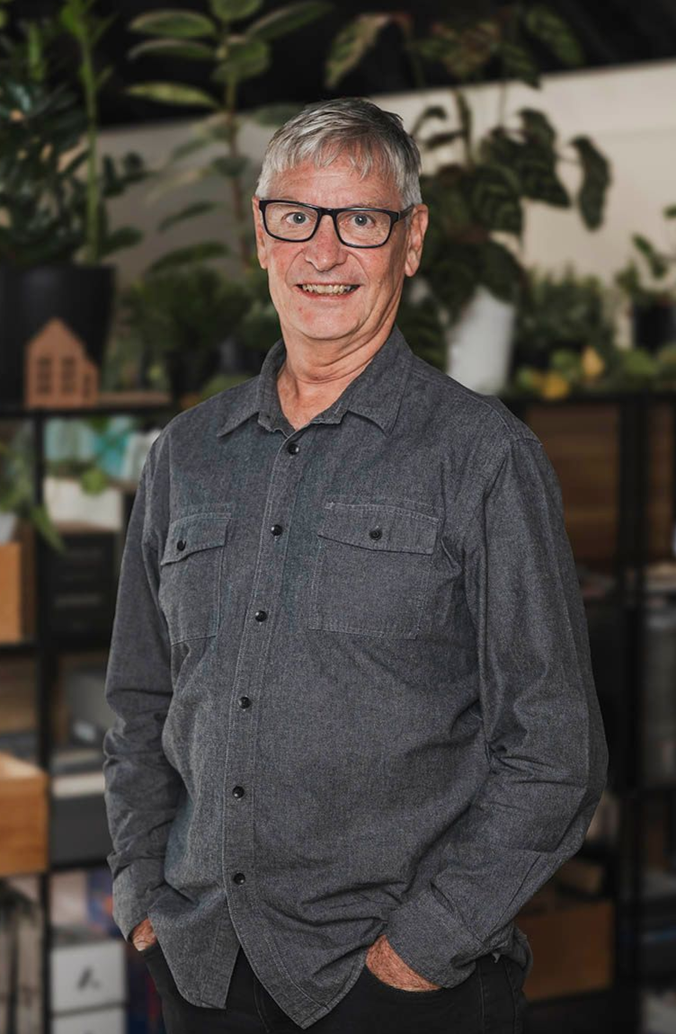 Smiling man in glasses and a gray shirt standing indoors against a leafy background