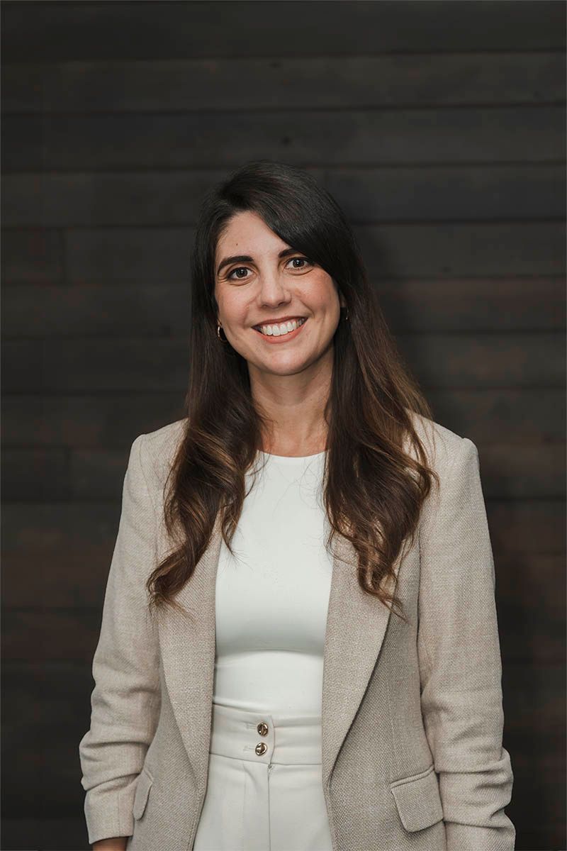 Smiling woman in a beige blazer and white top standing against a dark wood background