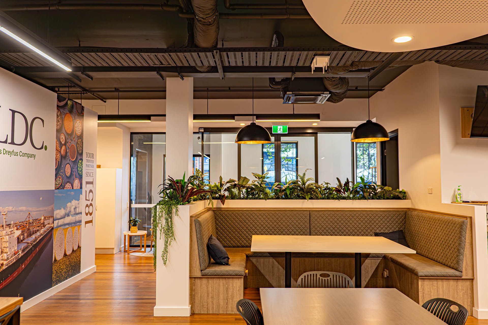 Modern office lounge with wooden booths, pendant lights, plants, and a wall mural in the background