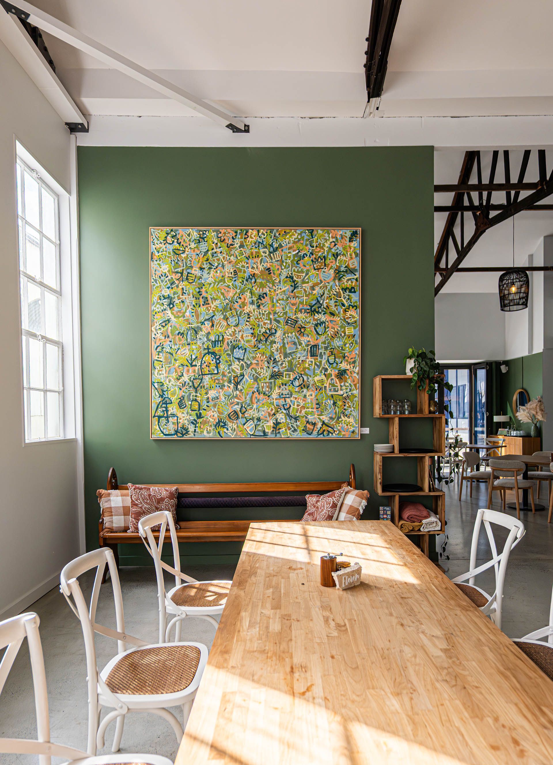 Bright café interior with a long wooden table, white chairs, green accent wall, and colorful artwork.