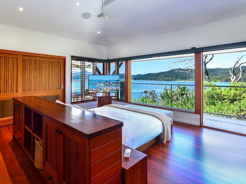 Bedroom with wooden furniture, ocean-view windows, and a tropical waterfront outside