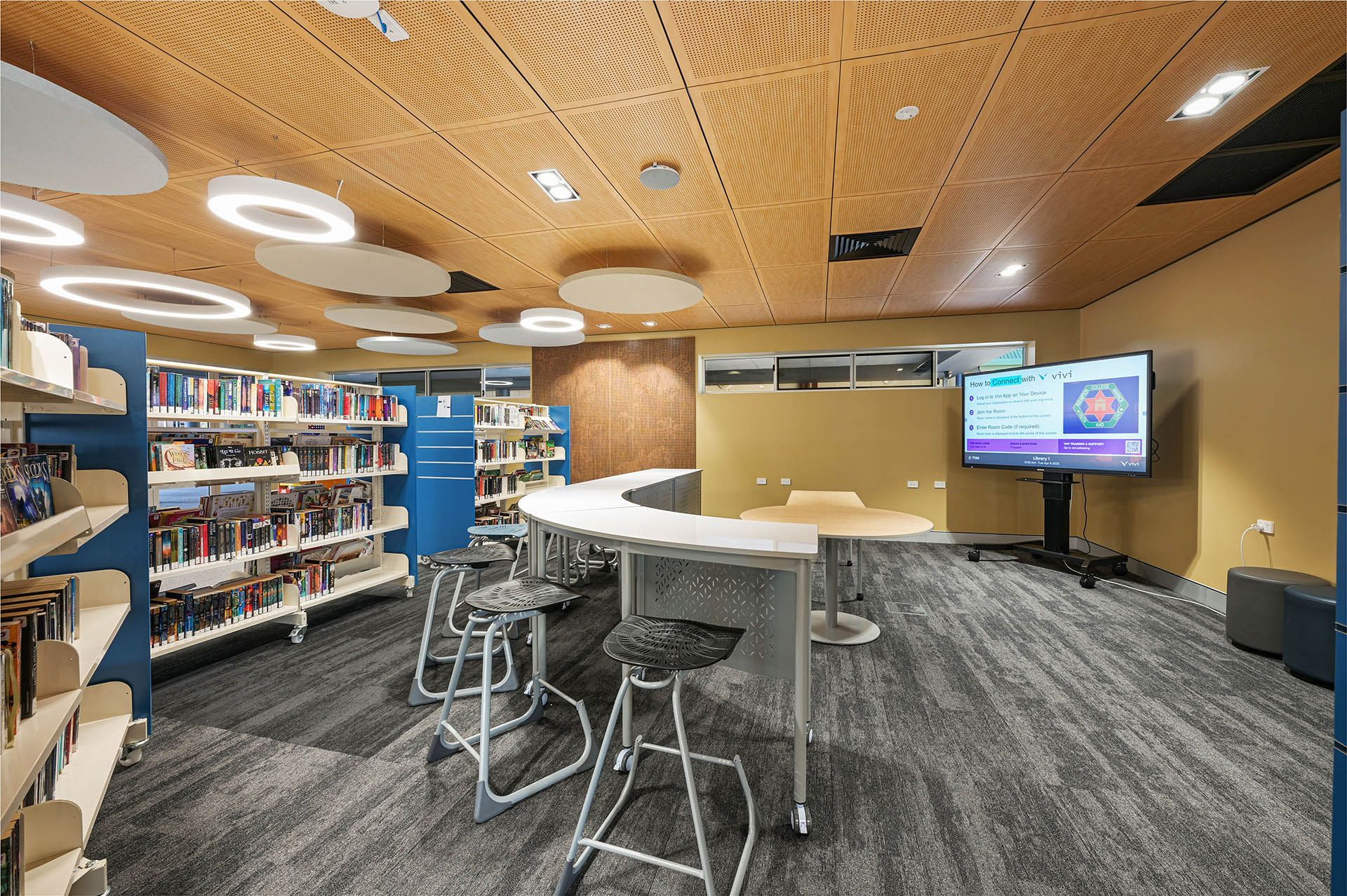 Library study area with bookshelves, round table, stools, and a wall-mounted TV on a carpeted floor.