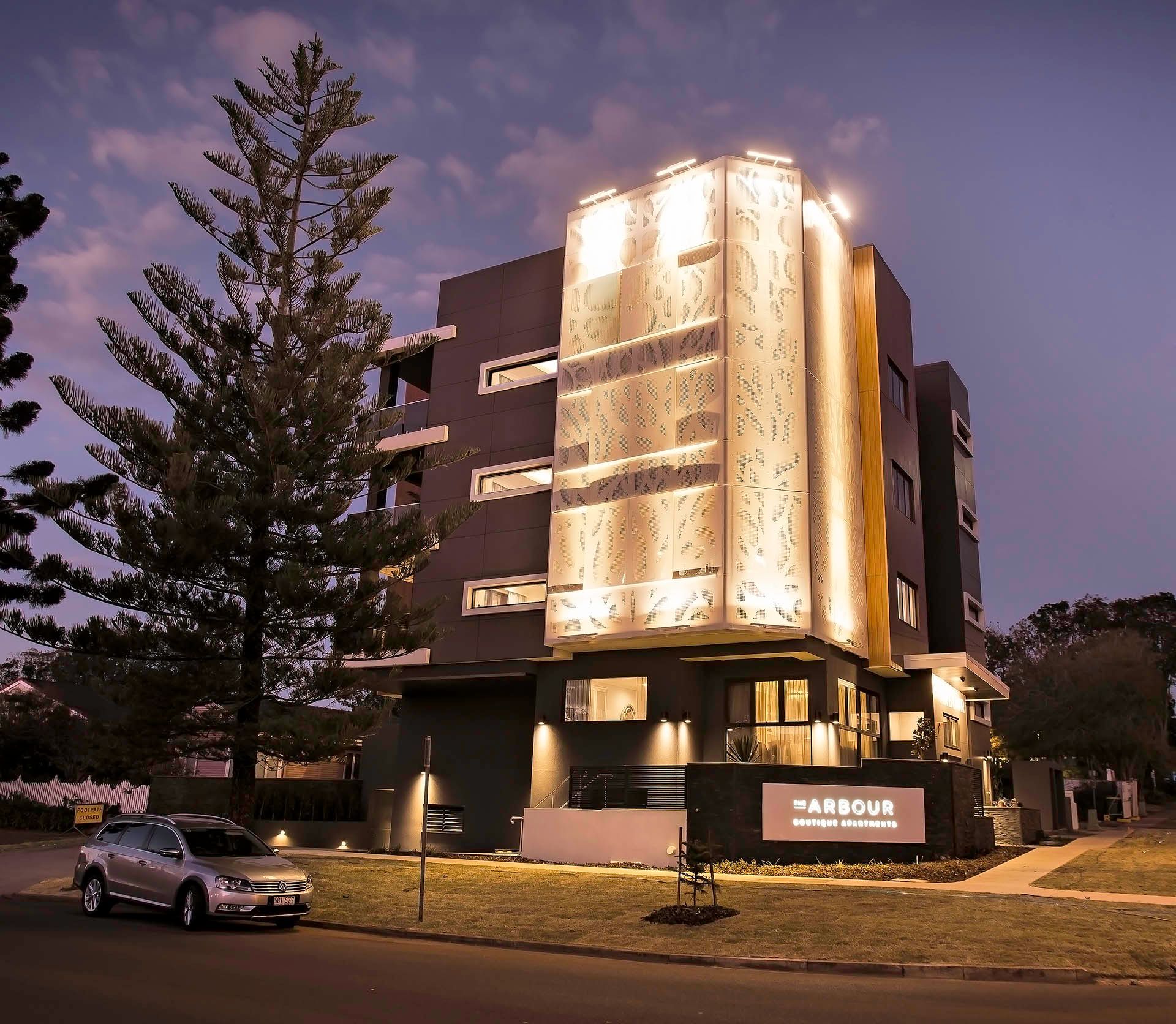 Modern multi-story hotel at dusk with glowing gold facade, parked cars, and a large tree in front