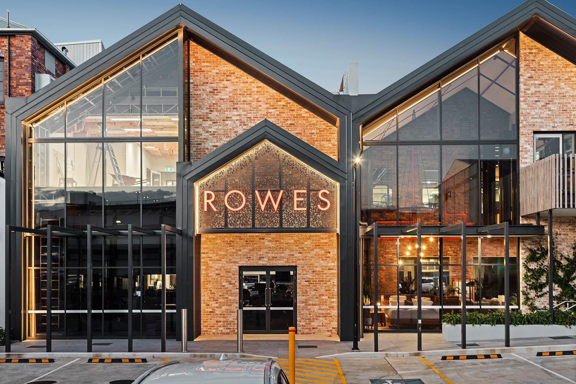 Modern brick storefront with large glass windows and illuminated “ROWES” sign at dusk
