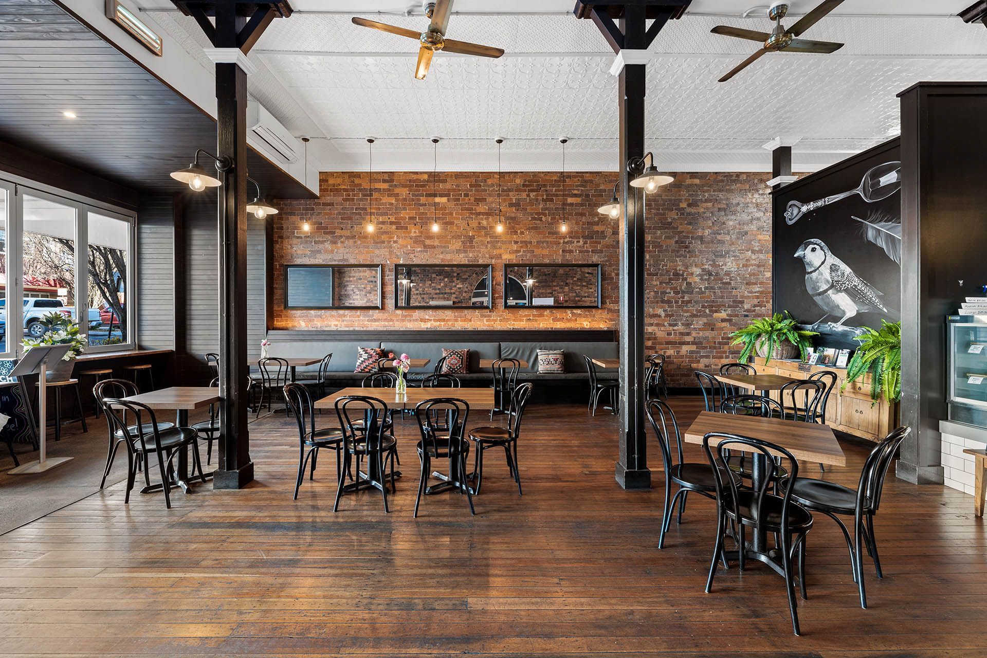 Bright café interior with brick wall, wooden tables, and pendant lights near large windows.