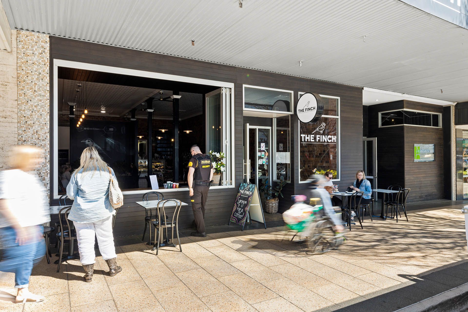 People sitting outside a café with black signage and large windows on a sunny sidewalk