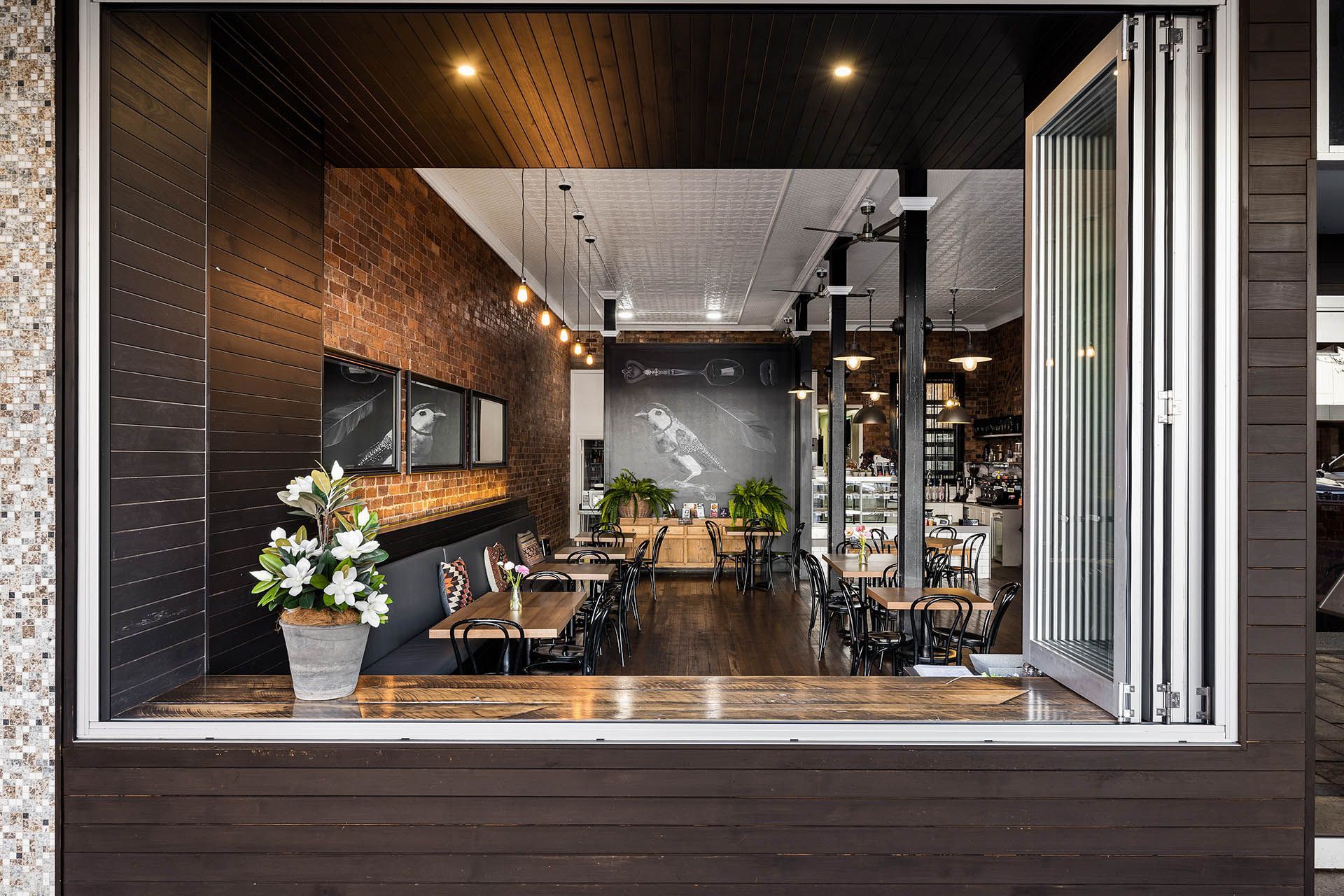 Modern café interior seen through a large open window, with tables, plants, and warm lighting.