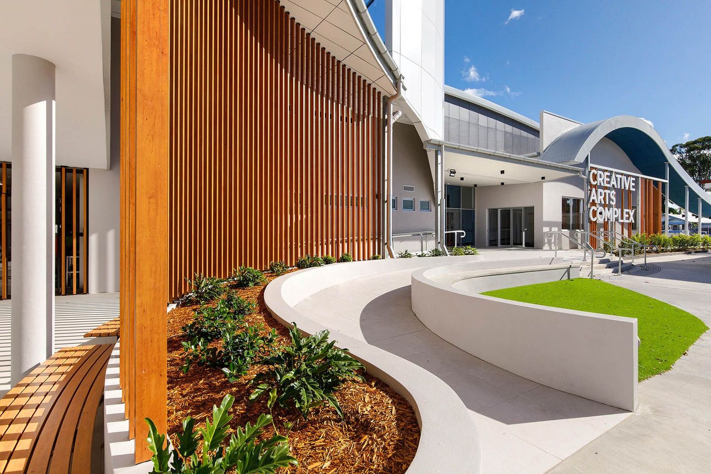 Modern building entrance with curved white walkway, wood slats, and landscaped greenery in bright sunlight